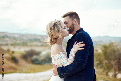 Couple in love hugs and kisses in fabulous mountains in nature. Girl in long white dress with bouquet of flowers in her hands, man in jacket. Wedding in nature, relationships and love