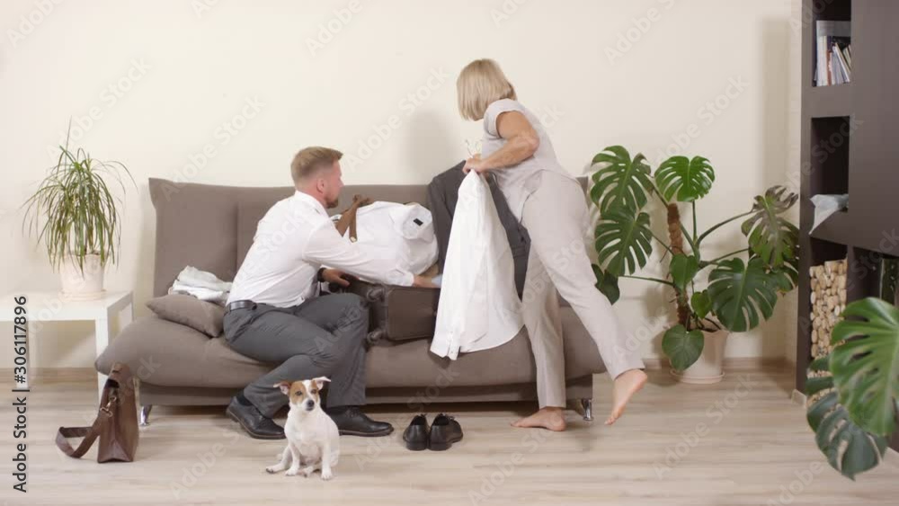 Tracking of cheerful blond woman walking into room carrying shirt and jacket, then helping her husband sitting on couch and packing suitcase for business trip Tracking of cheerful blond woman walking into room carrying shirt and jacket, then helping her husband sitting on couch and packing suitcase for business trip