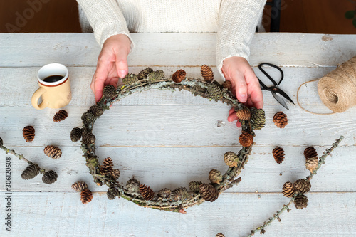 Fragment of a woman making a handmade larch wreath with cones as a symbol of natural Christmas on a white wooden table photo