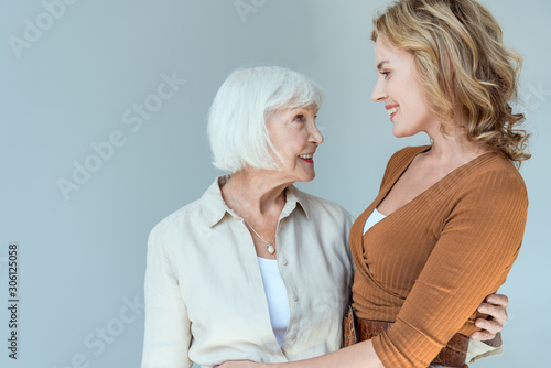smiling mother and daughter hugging and looking at each other isolated on grey