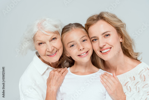 smiling mother and grandmother hugging granddaughter isolated on grey