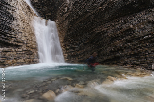woman alone practicing canyoning leaving the riverbed with a mountain waterfall behind her. Long exposition