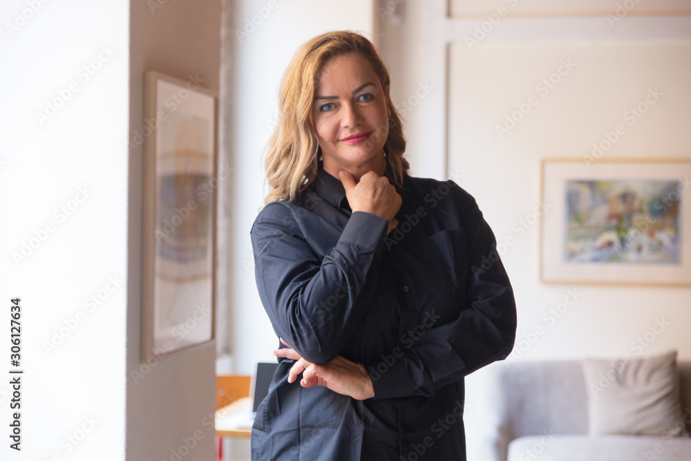 Positive middle aged lady posing in her living room at home. Happy Caucasian woman in casual standing for camera and smiling. Home female portrait concept Positive middle aged lady posing in her living room at home. Happy Caucasian woman in casual standing for camera and smiling. Home female portrait concept