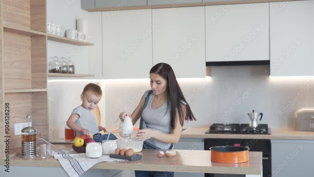 Cute little boy toddler and his mother is cooking in the modern kitchen together. Mother is mixing a dough in a bowl with a mixer and son is asking her about an apple. Cute little boy toddler and his mother is cooking in the modern kitchen together. Mother is mixing a dough in a bowl with a mixer and son is asking her about an apple.