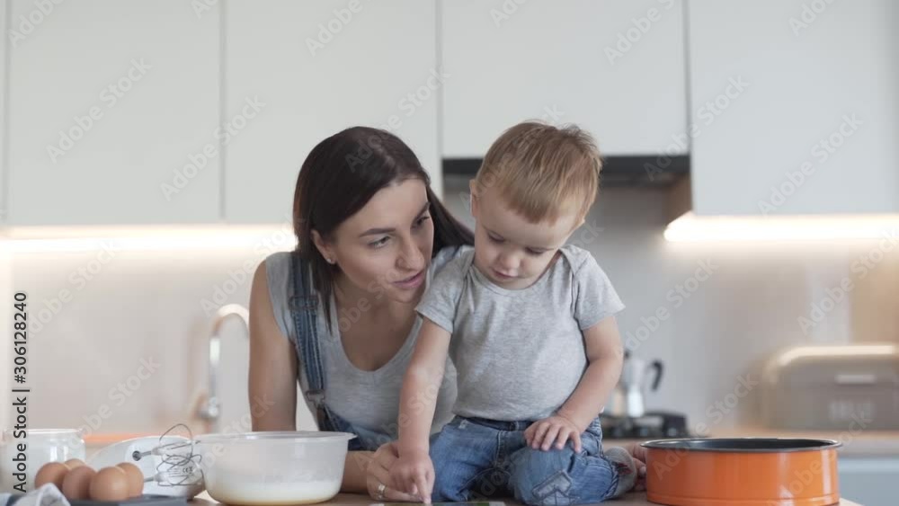 Young beautiful mother and her son are standing in a kitchen and watching a video on a phone. Woman is kissing her son in a cheek. Young beautiful mother and her son are standing in a kitchen and watching a video on a phone. Woman is kissing her son in a cheek.