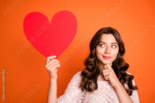 Portrait of minded girl hold big red paper card heart she get 14-february date think thoughts touch chin finger wear good looking outfit isolated over orange color background