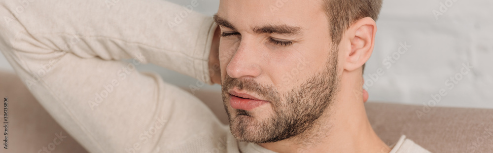 panoramic shot of man with closed eyes sitting on sofa and suffering from neck pain