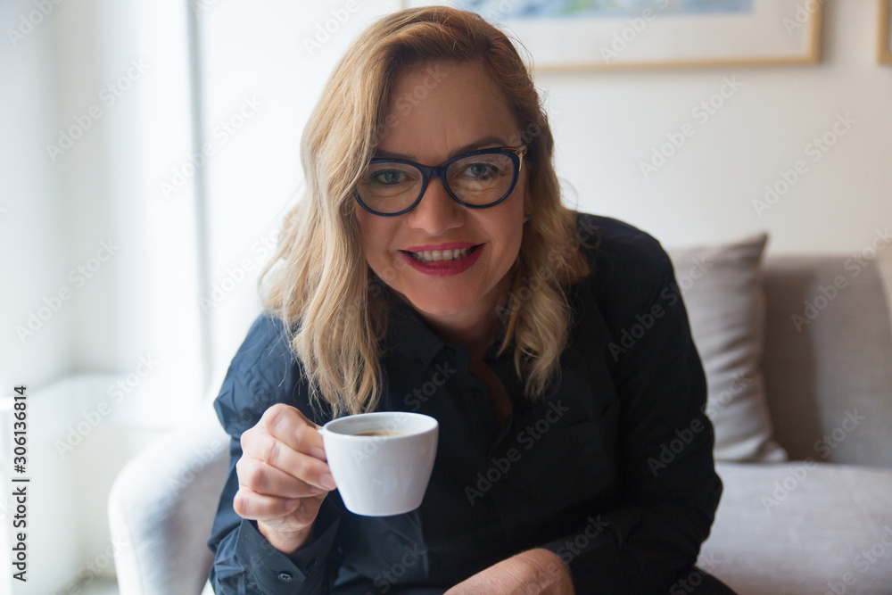 Joyful middle aged lady in glasses drinking coffee at home. Caucasian woman in casual holding cup, looking at camera. Morning or coffee break concept Joyful middle aged lady in glasses drinking coffee at home. Caucasian woman in casual holding cup, looking at camera. Morning or coffee break concept