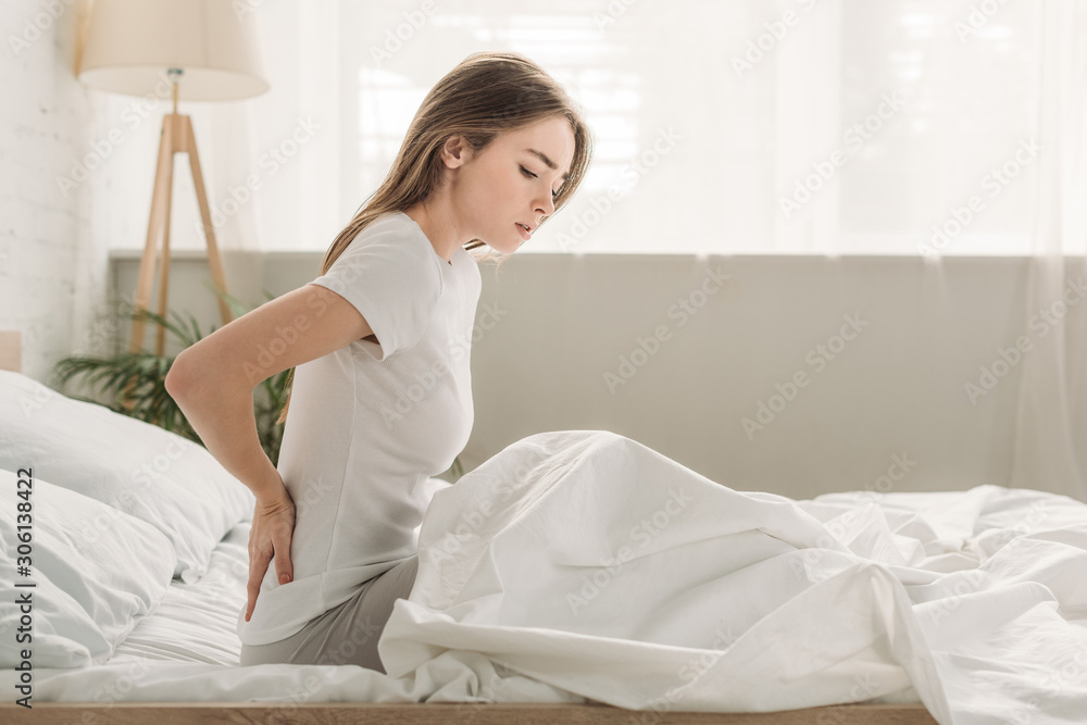 frowned girl touching loin while sitting in bed and suffering from pain frowned girl touching loin while sitting in bed and suffering from pain