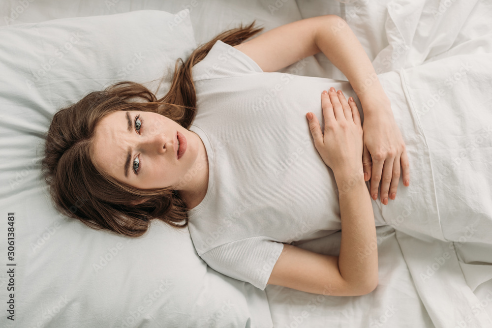 top view of upset girl lying in bed and suffering from stomach pain top view of upset girl lying in bed and suffering from stomach pain