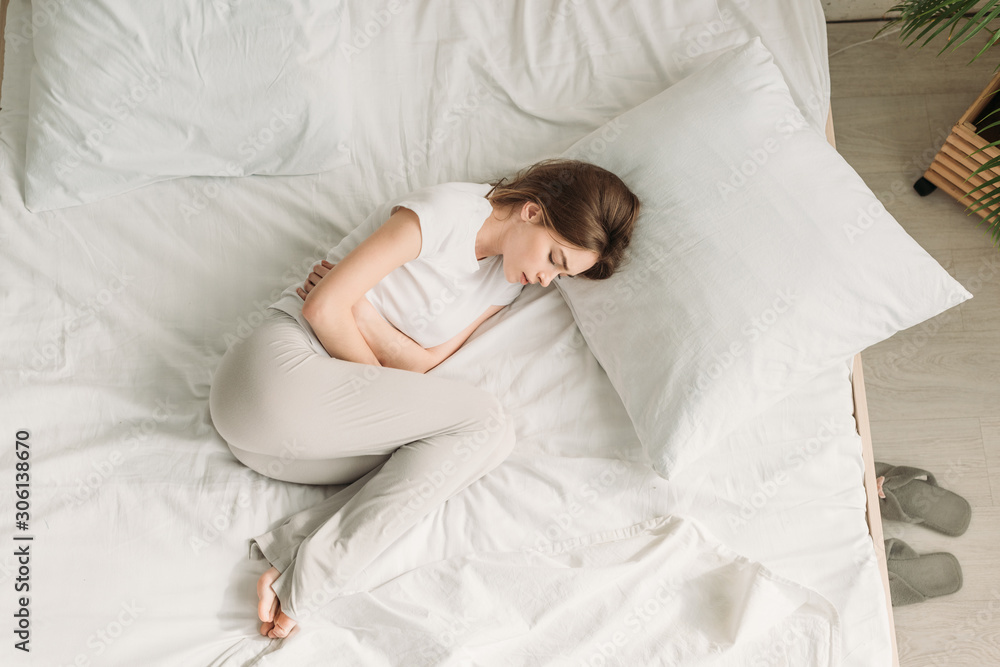 top view of young woman lying in bed and suffering from stomach pain top view of young woman lying in bed and suffering from stomach pain