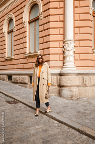 authentic street style portrait of an attractive woman wearing trench coat, sunglasses and slingback shoes, crossing the street.fashion outfit details perfect for autumn fall winter © mlasaimages