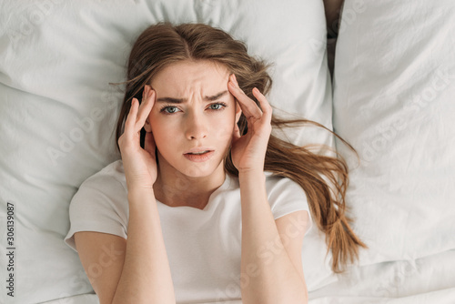 top view of young woman looking at camera while lying in bed and suffering from headache
