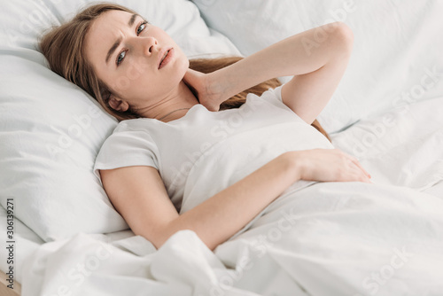 young woman looking at camera while lying in bed with closed eyes and suffering from neck pain