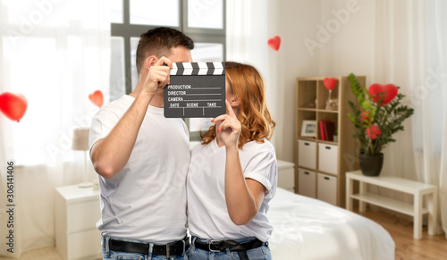 production, filmmaking and valentine's day concept - portrait of happy couple in white t-shirts with clapperboard over home bedroom decorated with heart shaped balloons background