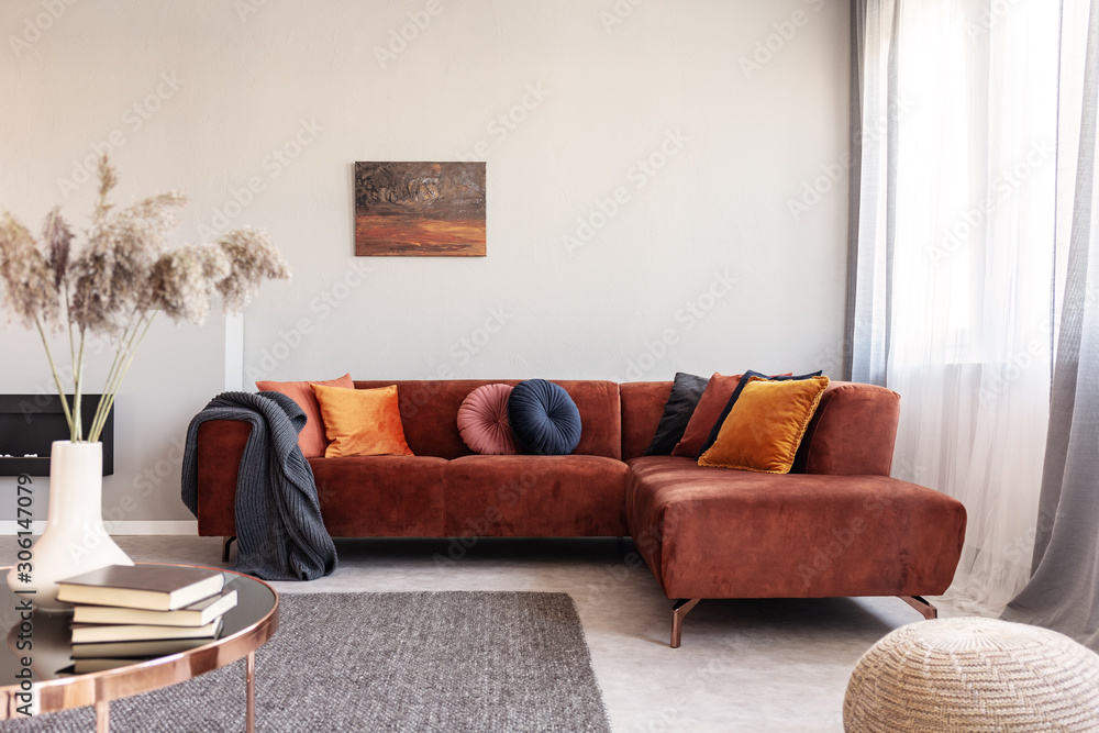 Real photo of warm color cushions on a red couch standing next to the window in cozy living room interior - obrazy, fototapety, plakaty 
