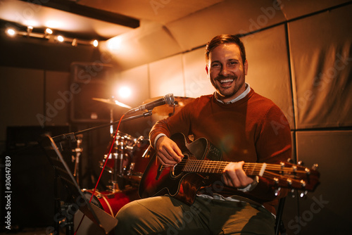 Portrait of a happy musician playing guitar and smiling at camera, in recording studio.
