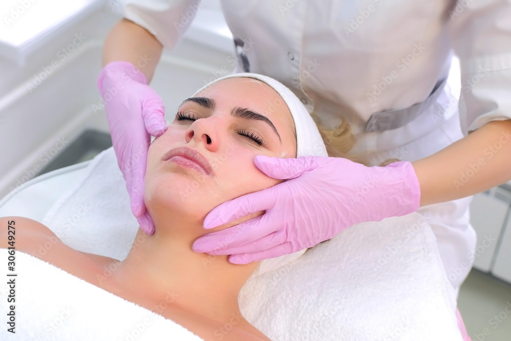 Cosmetologist in gloves washes client's face, neck and chest with gel. Portrait of young woman, closeup view. Beautician making beauty procedure to patient relaxing in clinic. Beauty industry concept.