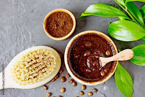 Coffee peeling. Coffee scrub in a wooden bowl with a wooden spoon on a dark background. Nearby is a bowl with coffee grounds, coffee grains, body brush and green flower leaves. Close-up 