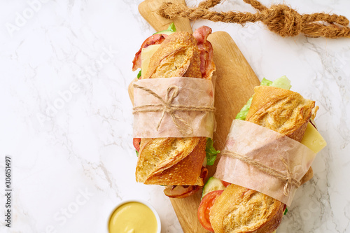 Two fresh big baguette sandwiches with bacon, chedder cheese, mustard, lettuce and vegetables on cutting board on marble background photo