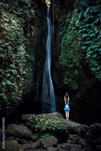 Woman standing on the rock, practicing yoga. Young woman raising arms with namaste mudra near waterfall. Leke Leke waterfall, Bali. View from back.