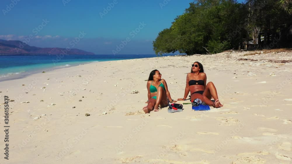 Beautiful, Sexy Women Tourists In Bikinis Laying On The White Sand Beach Of Indonesia Enjoying The Sun With Waves, Trees And Mountains In The Background - Wide Shot