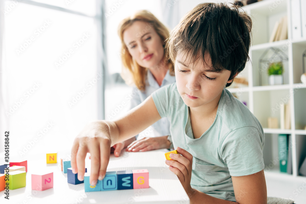 selective focus of kid with dyslexia playing with building blocks and child psychologist looking at it on background - obrazy, fototapety, plakaty 
