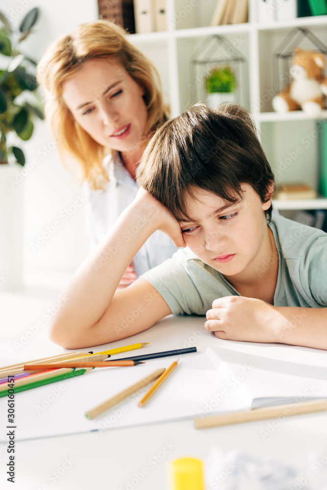 selective focus of sad kid with dyslexia sitting at table and child psychologist looking at him on background selective focus of sad kid with dyslexia sitting at table and child psychologist looking at him on background