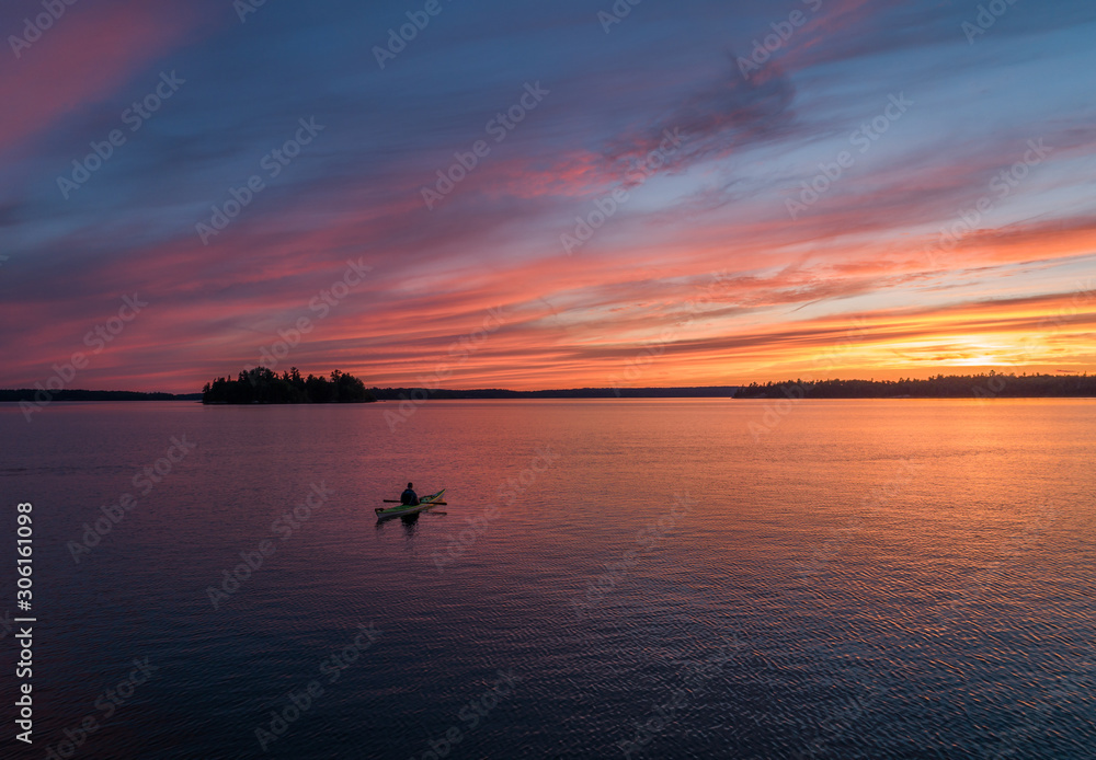 A lone kayaker contemplates the meaning of life while in a meditative moment out in the middle of a calm lake under a colorful northern sky.