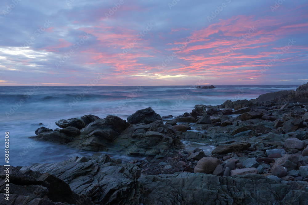 Motion-blurred waves crashing on a rocky shoreline under a red sunset