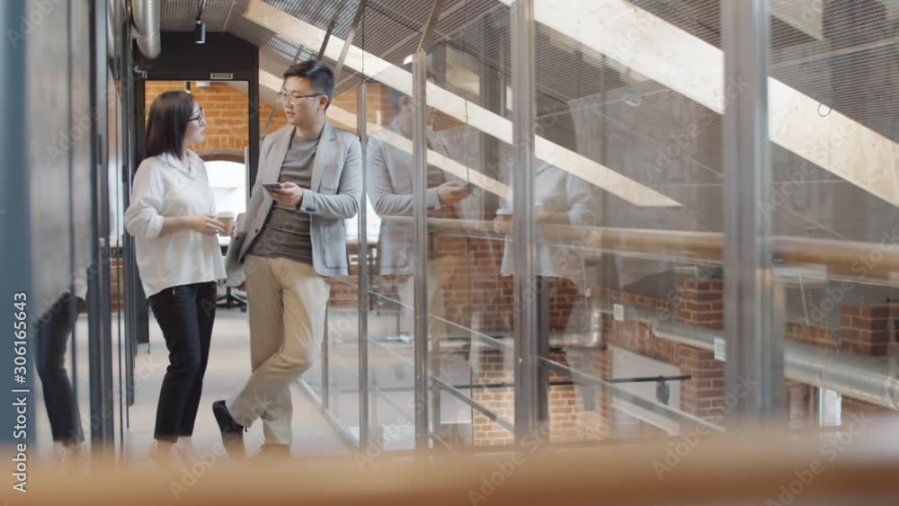 Wide shot of young Asian businessman leaning on glass wall and businesswoman standing together in office, looking at smartphone screen and talking Wide shot of young Asian businessman leaning on glass wall and businesswoman standing together in office, looking at smartphone screen and talking