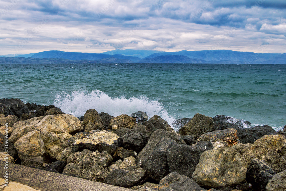 Seascape at stormy weather – waves at sea, large rocks on the shore, dark clouds on the sky and mountains on the horizon