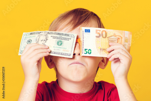 Joyful kid covering eyes with dollar and euro bill over yellow. Happy little boy enjoying his income