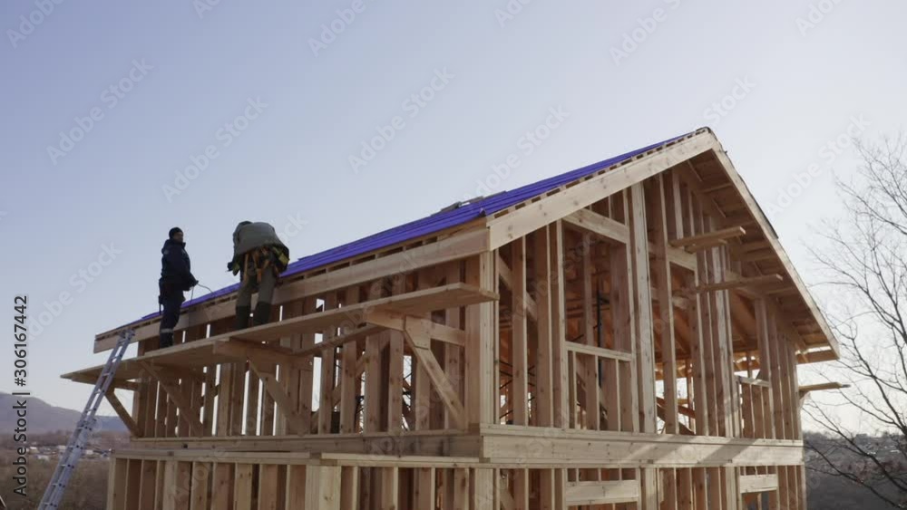Two builders are lying soft roof working at height. Aerial panoramic view of wooden frame house under construction Two builders are lying soft roof working at height. Aerial panoramic view of wooden frame house under construction