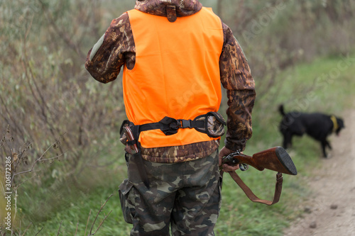 A man with a gun in his hands and an orange vest on a pheasant hunt in a wooded area in cloudy weather. Hunter with dogs in search of game.