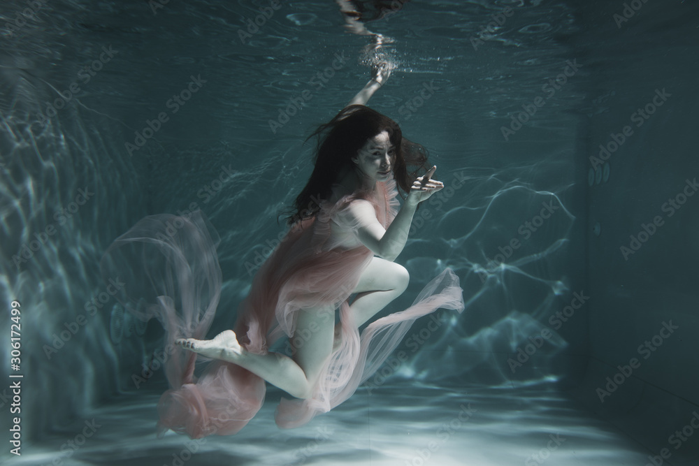 Fototapeta A girl with long dark hair swims underwater in a pink dress and with a crown on her head, like an underwater queen. Fairy tale suitable for advertising