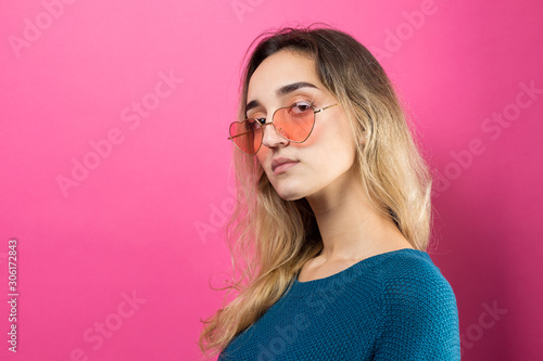 Romantic blonde-haired girl in trendy heart glasses posing with shy smile. Studio close-up portrait on a pink background.