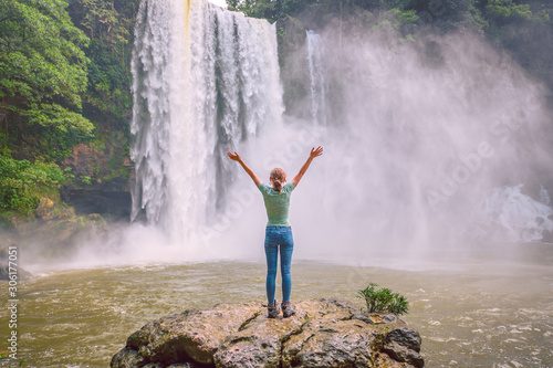 Young blond woman with arms raised standing on a rock watching the landscape with a waterfall in the middle of the jungle