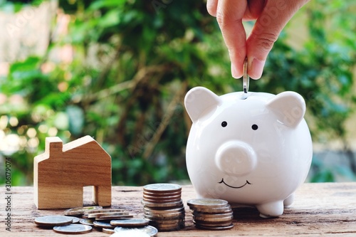 Money for housing. Putting each coin into a piggy bank. Wooden house model and money with greenery background.