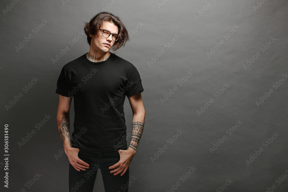 Hipster handsome male model with glasses wearing black blank t-shirt and black jeans with space for your logo or design in casual urban style - obrazy, fototapety, plakaty 