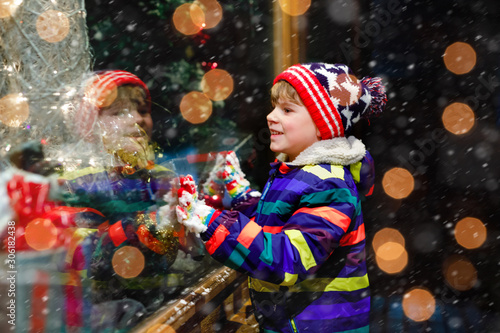 Cute little healthy school kid boy on Christmas market. Funny happy child in fashion winter clothes making window shopping decorated with gifts, xmas tree. Snow falling down, snowfall