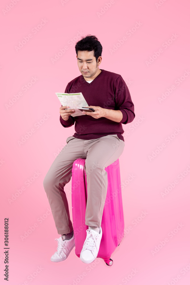 Happy Asian man sitting on luggage, holdig map, passport on pink background. Happy Asian man sitting on luggage, holdig map, passport on pink background.