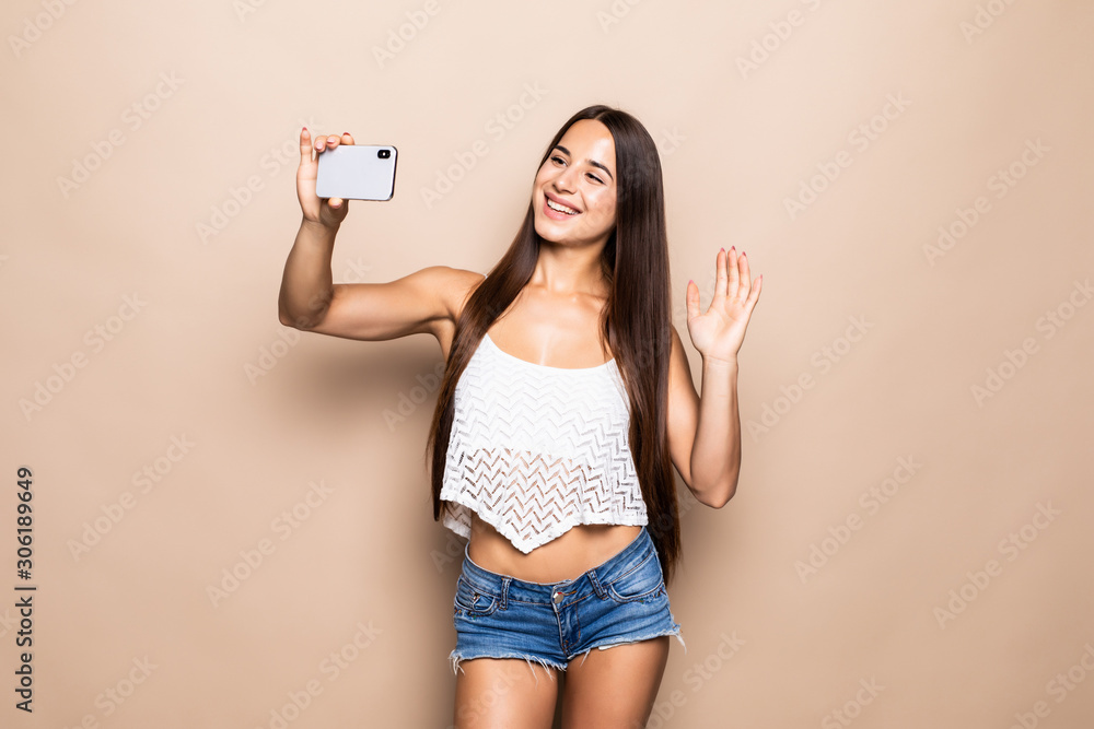 Portrait of a pretty young girl taking selfie with mobile phone and waving isolated over beige background Portrait of a pretty young girl taking selfie with mobile phone and waving isolated over beige background