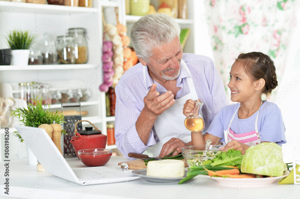 Portrait of senior man with granddaughter looking at laptop while preparing dinner in kitchen Portrait of senior man with granddaughter looking at laptop while preparing dinner in kitchen