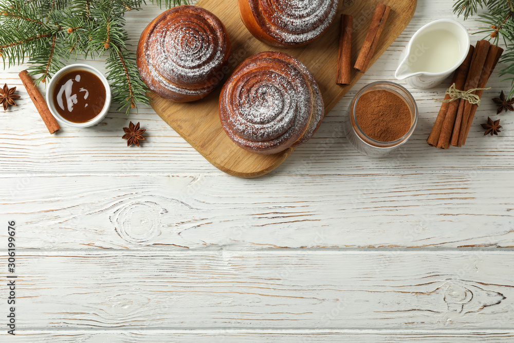 Cinnamon rolls, caramel, milk and fir branches on white wooden background, top view