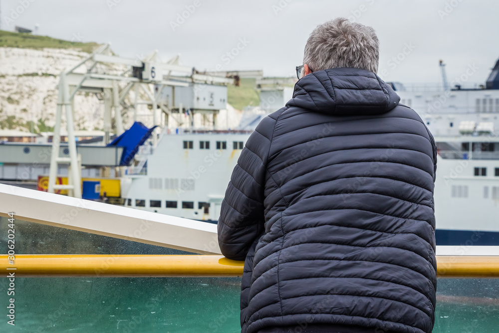 Dover, England, 15/05/2019 Man looking over the bannister of a P&O ferry at the coast of dover with a coat grey hair old glasses and beard vaping on a ship boat with coat on cold weather