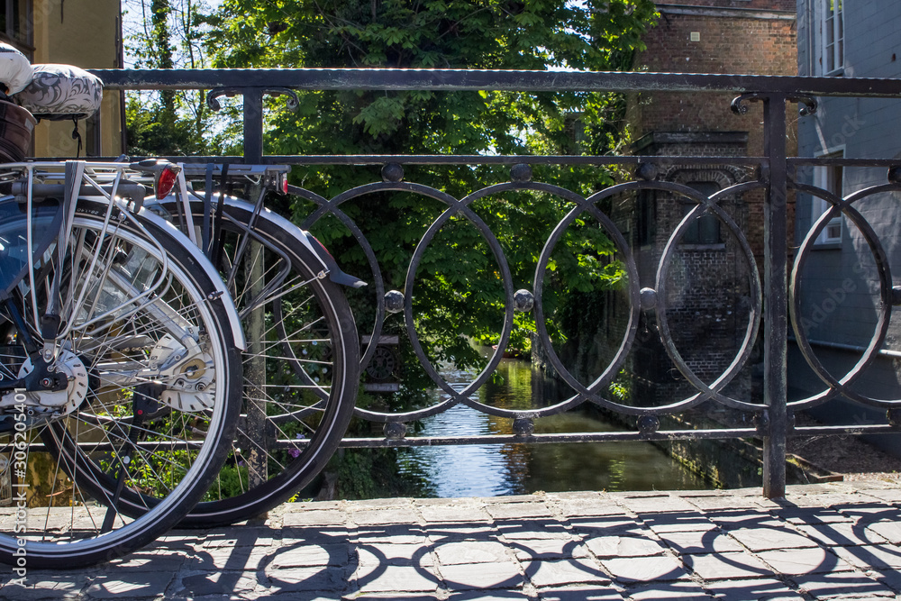 Two bicycles against a metal wall fence barrier on a bridge in europe over water with reflection of the sky and greenery trees grass brushes close up on the wheels of the bikes and french belguim vibe