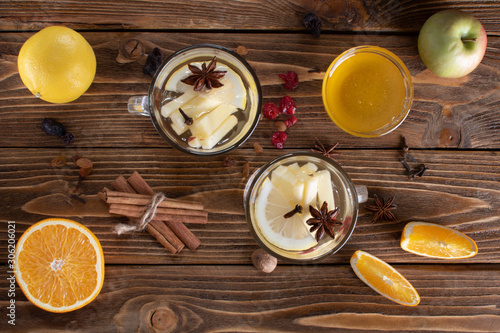 Top view of two glasses with white mulled wine with a slice of orange, lemon, apple, anise and a stick of chicken, with scattered dried fruits on a wooden table.