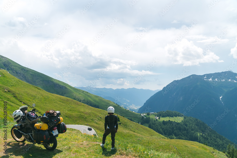 Biker woman wearing protective turtle jacket. standing near his tourer motorcycle on the green grass. Looking into distance. Alpine mountains.