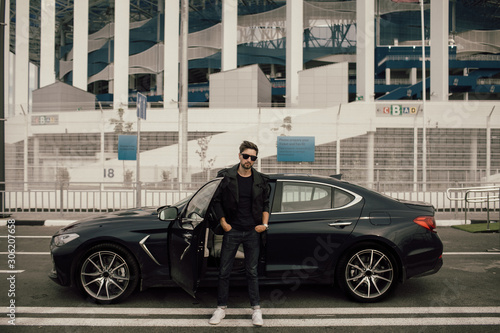 a man in sunglasses and a leather jacket stands near a sports car